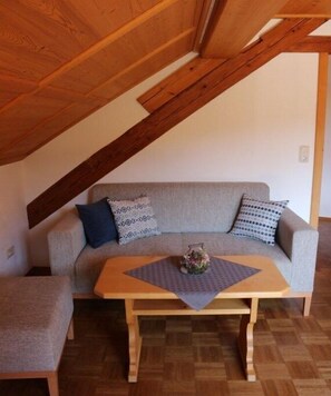 Ein Loft-Zimmer mit Holzbalkendecke, einem grauen Sofa mit gemusterten Kissen und einem kleinen Tisch mit einer Vase.