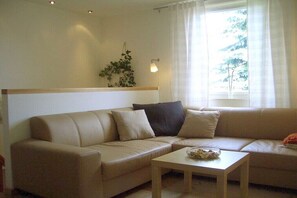 Ein Wohnzimmer mit einem beigen Sofa, einem kleinen Tisch, einer Pflanze und einem Fenster mit Vorhängen.