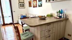 Eine kleine Küche mit Essbereich, einer Couch und einem Tisch mit Schüssel und Tellern. An der Wand befinden sich gerahmte Bilder und ein Regal mit Flaschen.