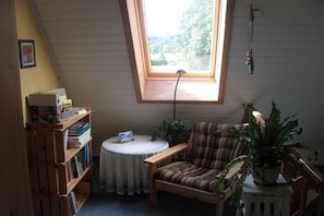 Ein Zimmer mit schrägverdeckter Decke, einem Fenster, einem Bücherregal, einem runden Tisch, einem gestreiften Sessel und einer Topfpflanze.