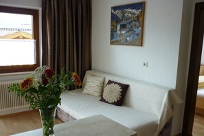 Ein Wohnzimmer mit einem weißen Sofa, einer Blumenvase, einem Bild an der Wand und einem Fenster mit Vorhängen.