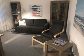 Ein Wohnzimmer mit einem dunkelgrauen Sofa, einem Holzschaukelstuhl, einem kleinen Tisch mit einer Zeitschrift, einem Bücherregal und einer Wandillustration von einem Fisch.