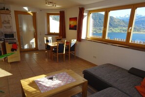 Ein gemütlicher Wohnbereich mit Essplatz, einer Couch, einem Tisch mit Unterlage und Blick auf einen See durch große Fenster.