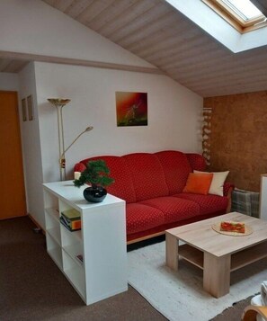 Ein Wohnzimmer mit einem roten Sofa, einem kleinen Tisch mit Pflanze, einer stehenden Lampe und einem Bild an der Wand.