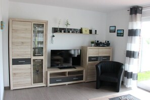 Ein Wohnzimmer mit Fernseher, Holzschränken, einem schwarzen Sessel und einem Fenster mit Vorhängen.