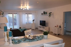 Ein Wohnzimmer mit Fernseher, weißen Möbeln, einem Esstisch mit Kerzen und einem Fenster mit Vorhängen.
