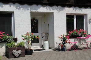 Ein Gebäude mit weißer Fassade, einer Tür mit Blumenkranz und Topfpflanzen mit rosa Blüten.