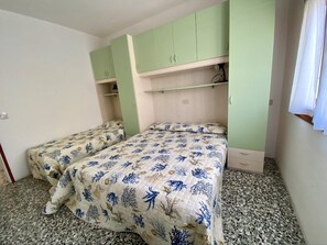 Ein Zimmer mit zwei Betten, grünen Schränken, einem kleinen Regal und einem Fenster.