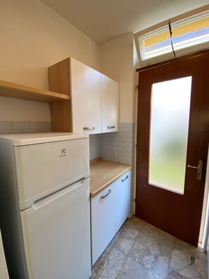 Eine kleine Küche mit weißen Schränken, einem weißen Kühlschrank, einer Holz Arbeitsplatte und einer Milchglas-Tür.