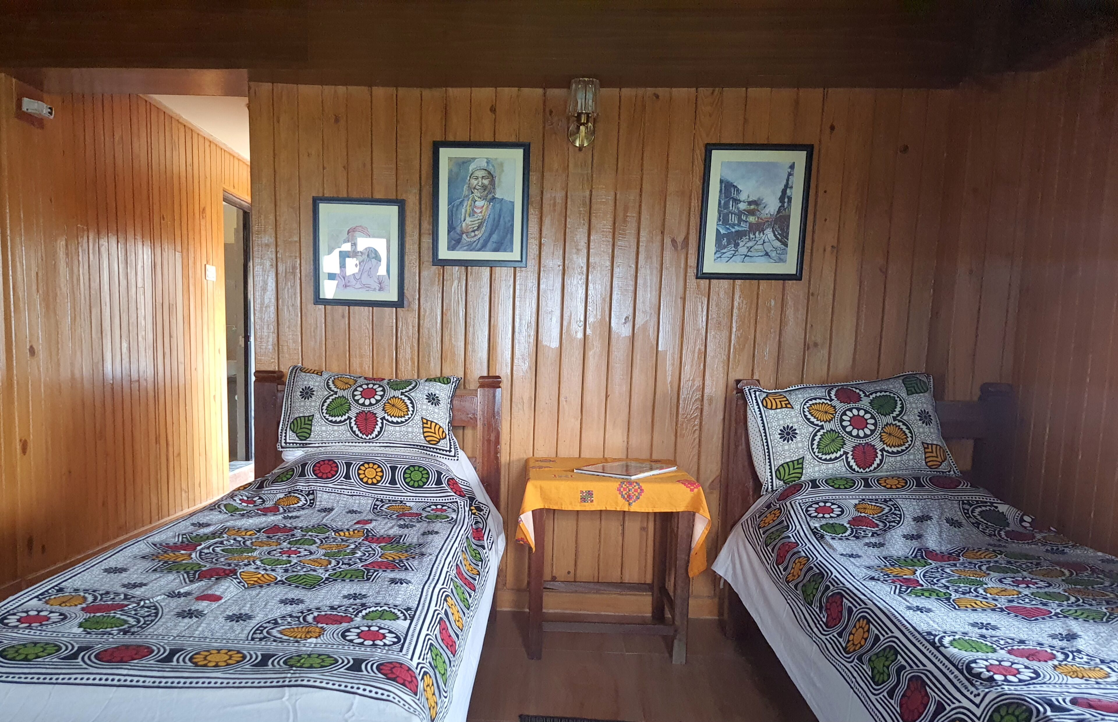 Une pièce aux murs lambrissés, avec deux lits, chacun recouvert d’une couvre-lit aux motifs colorés. Des tableaux encadrés ornent le mur, et une petite table est recouverte d’une nappe jaune.