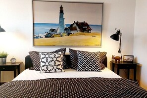 Ein Bett mit Punkteoptik, zwei gemusterte Kissen, ein Gemälde eines Leuchtturms, eine Lampe und ein kleiner Tisch mit Pflanze und Bilderrahmen.