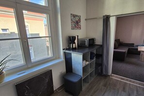 Ein kompaktes Zimmer mit Fenster, einer kleinen Küchenzeile inklusive Kaffeemaschine und Mikrowelle, einer Topfpflanze und einer Schiebetür zu einem Außenbereich.