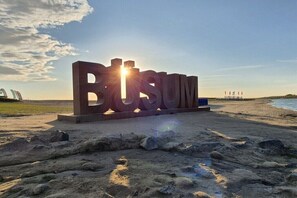 Ein großes Hinweisschild mit dem Schriftzug 'BUSUM' am Strand mit Sand, Steinen und einem Gewässer im Hintergrund.
