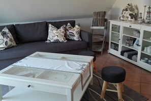Ein Wohnzimmer mit grauem Sofa, gemusterten Kissen, einem weißen Schrank mit Glastüren und einem kleinen Hocker.