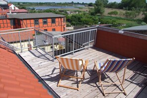 A wooden deck with two striped beach chairs, a metal railing, and a view of a lake and buildings in the background. - Holiday apartment with 1 bedroom (Röbel \/ Müritz)