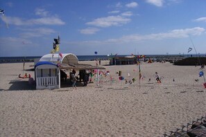 Ein Strand mit einer kleinen Laube, einem Pier im Hintergrund und Menschen bei verschiedenen Aktivitäten.
