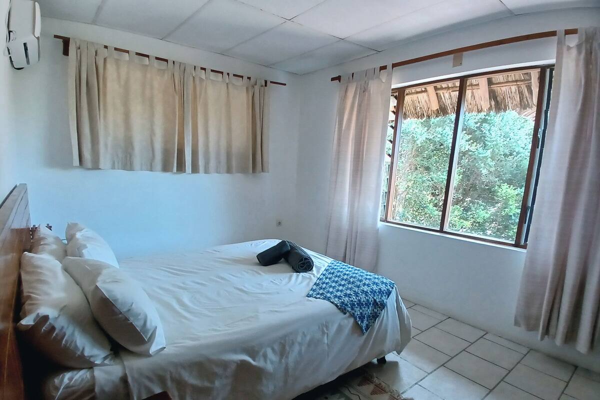 A neatly made bed with pillows, a blue patterned blanket, and a rolled-up towel. There is a window with curtains, a wooden headboard, and a wall-mounted air conditioning unit.