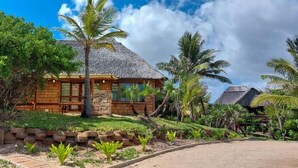 Une maison en bois avec un toit de chaume, entourée d’une végétation luxuriante et de palmiers.