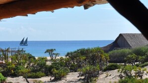 Une vue sur la plage avec une structure à toit de chaume, des palmiers et un voilier sur l’eau.
