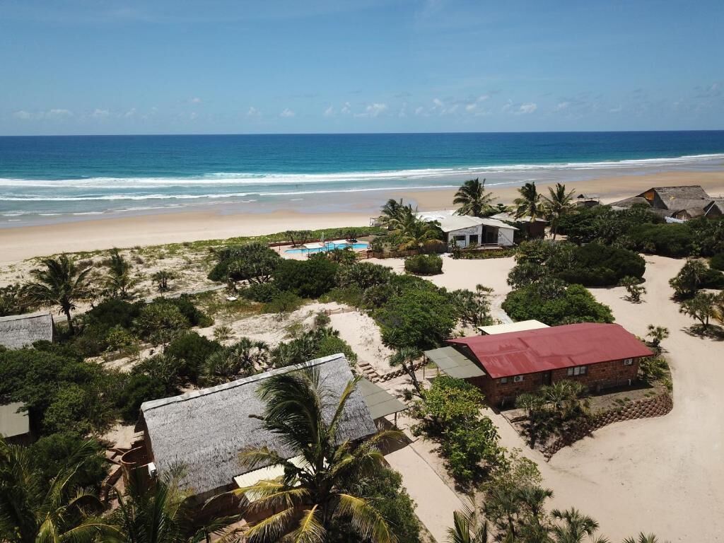 Vista aérea de una propiedad frente a la playa con piscina, cabañas de playa y un edificio de techo rojo.