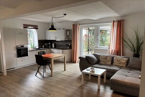 Eine moderne Küche mit weißen Schränken, einem Esstisch und einer Couch in einem Wohnbereich mit großem Fenster.