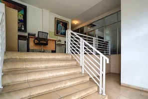 Un escalier recouvert de carreaux beiges et doté de rampes blanches mène à une pièce où se trouvent un bureau, une chaise et un tableau encadré accroché au mur.