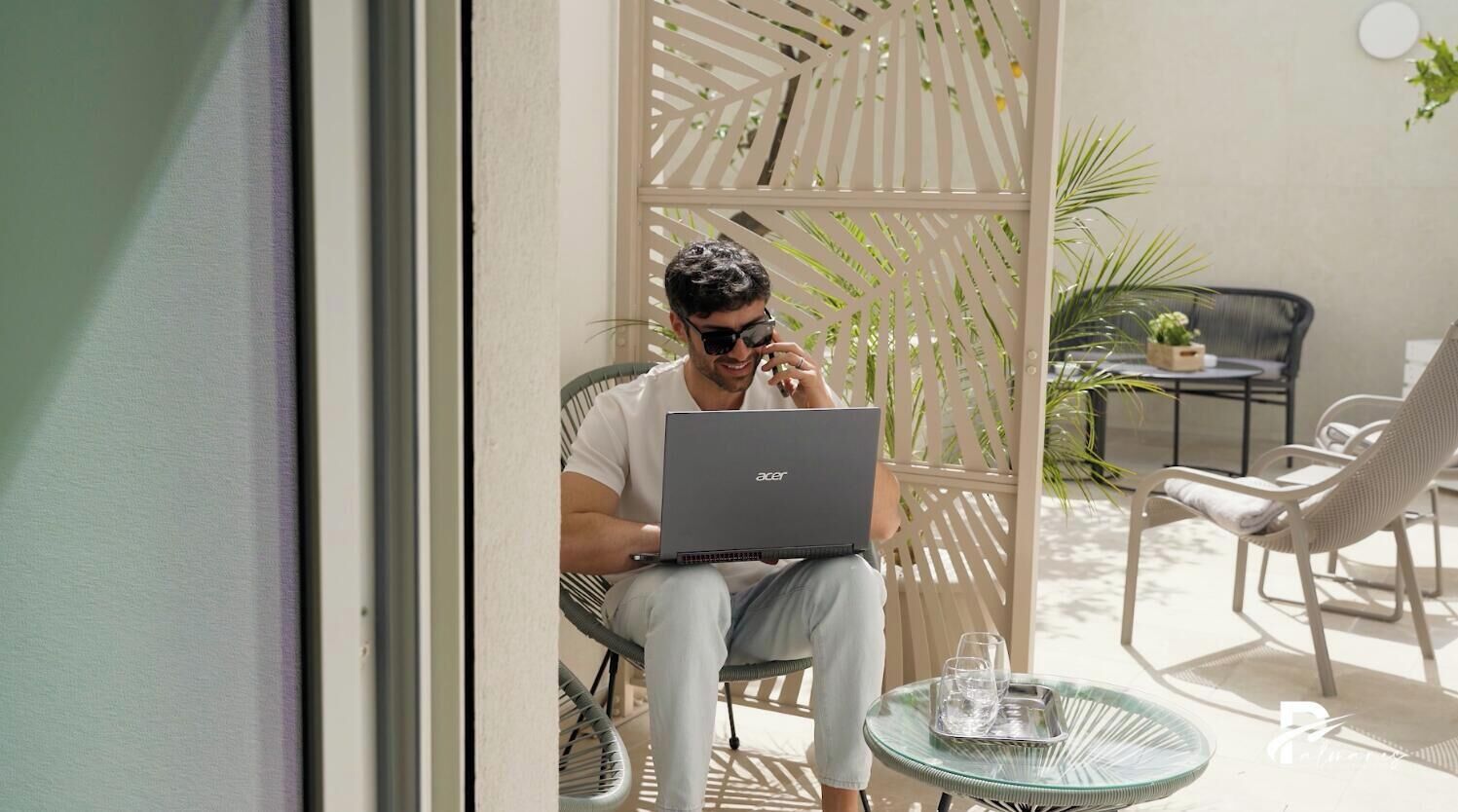 Eine Person sitzt auf einem Stuhl, auf einem kleinen Tisch stehen ein Laptop, ein Glas Wasser und ein weiteres Glas. Im Raum ist eine weiße Trennwand und weitere Stühle zu sehen.