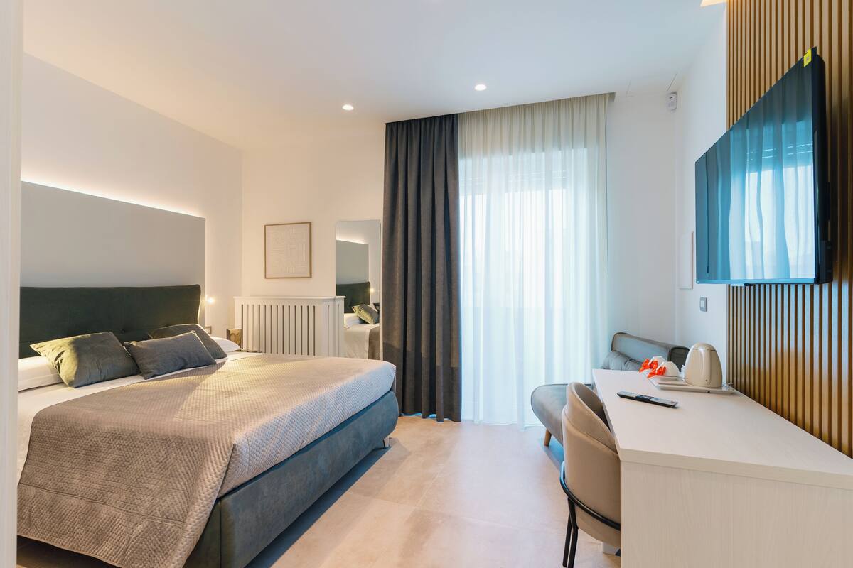 A modern hotel room with a large bed, a desk, a chair, a flat-screen TV, and a window with curtains.