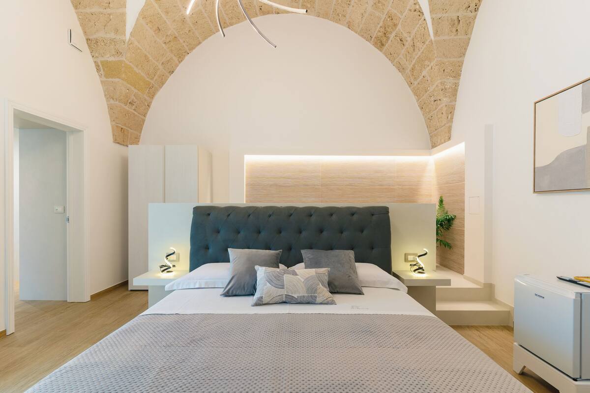 A bedroom with a tufted headboard, a bed with patterned bedding, bedside lamps, and a small table with a plant.