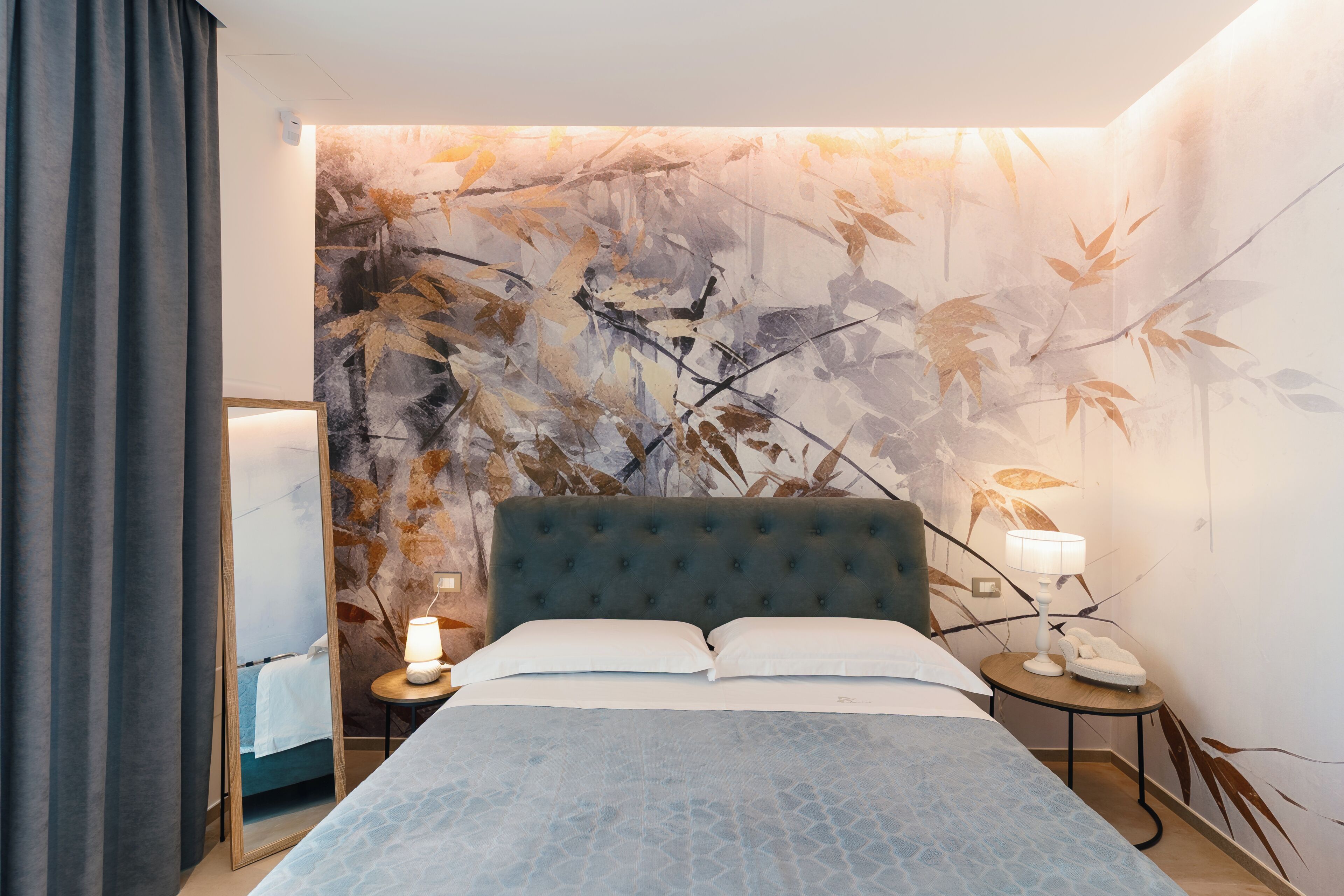 Un dormitorio con cabecera capitonada, cama con edredón estampado, mesita de noche con lámpara y un mural en la pared.