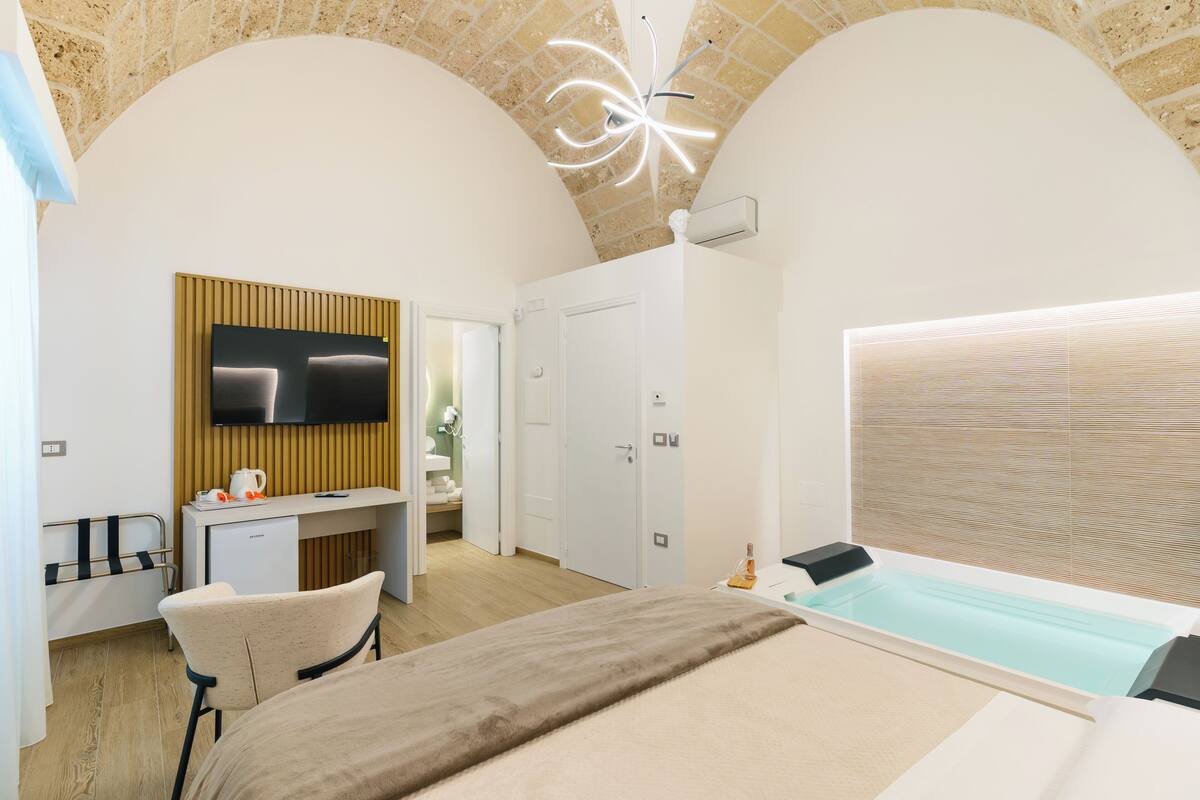 A modern hotel room with a large bed, a TV mounted on a wooden panel, a desk with a chair, and a bathtub.