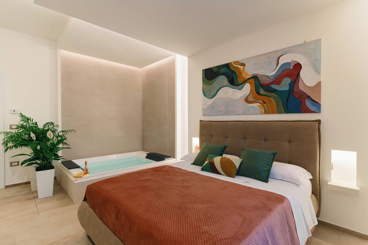 A modern hotel room with a large bed, a separate shower area, a potted plant, and a colorful abstract painting on the wall.