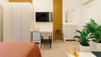 Una habitación de hotel moderna con cama, escritorio, silla, televisión, radiador, plantas en macetas y una botella en la mesita de noche.