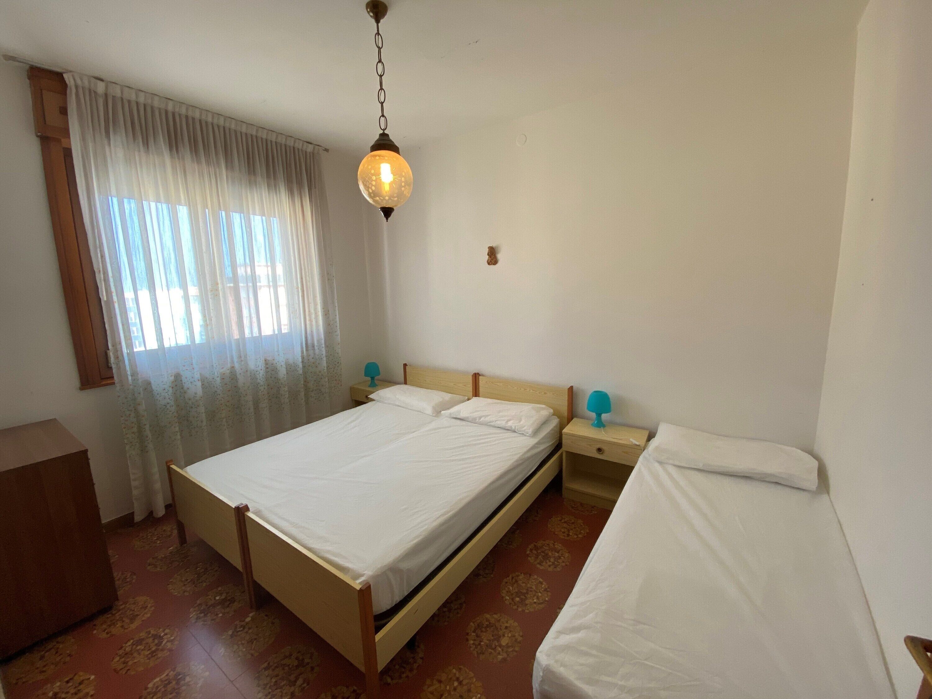 Une chambre avec un lit simple, une table de chevet, une lampe, une tête de lit en bois, une fenêtre avec des rideaux légers et un luminaire suspendu.