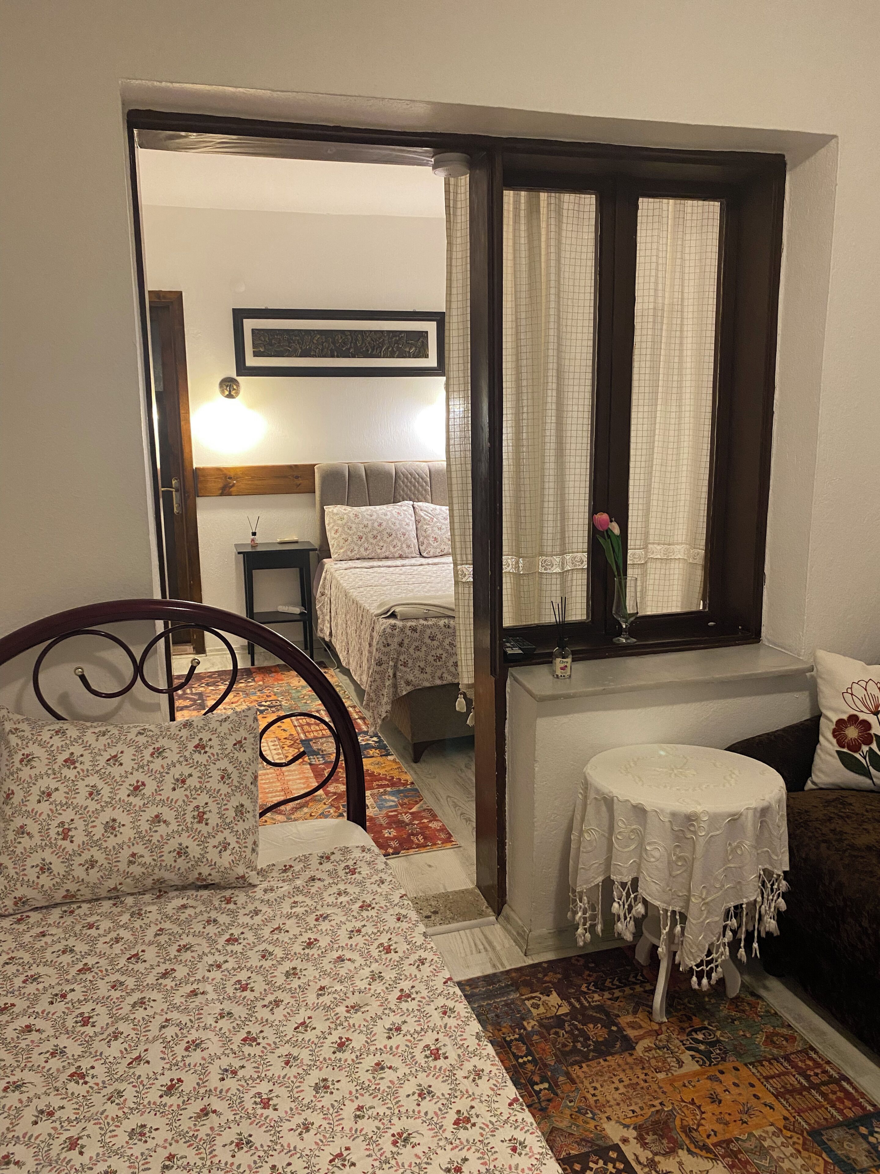 A bedroom with a bed, a bedside table, a small round table, a window with curtains, and a door leading to another room.