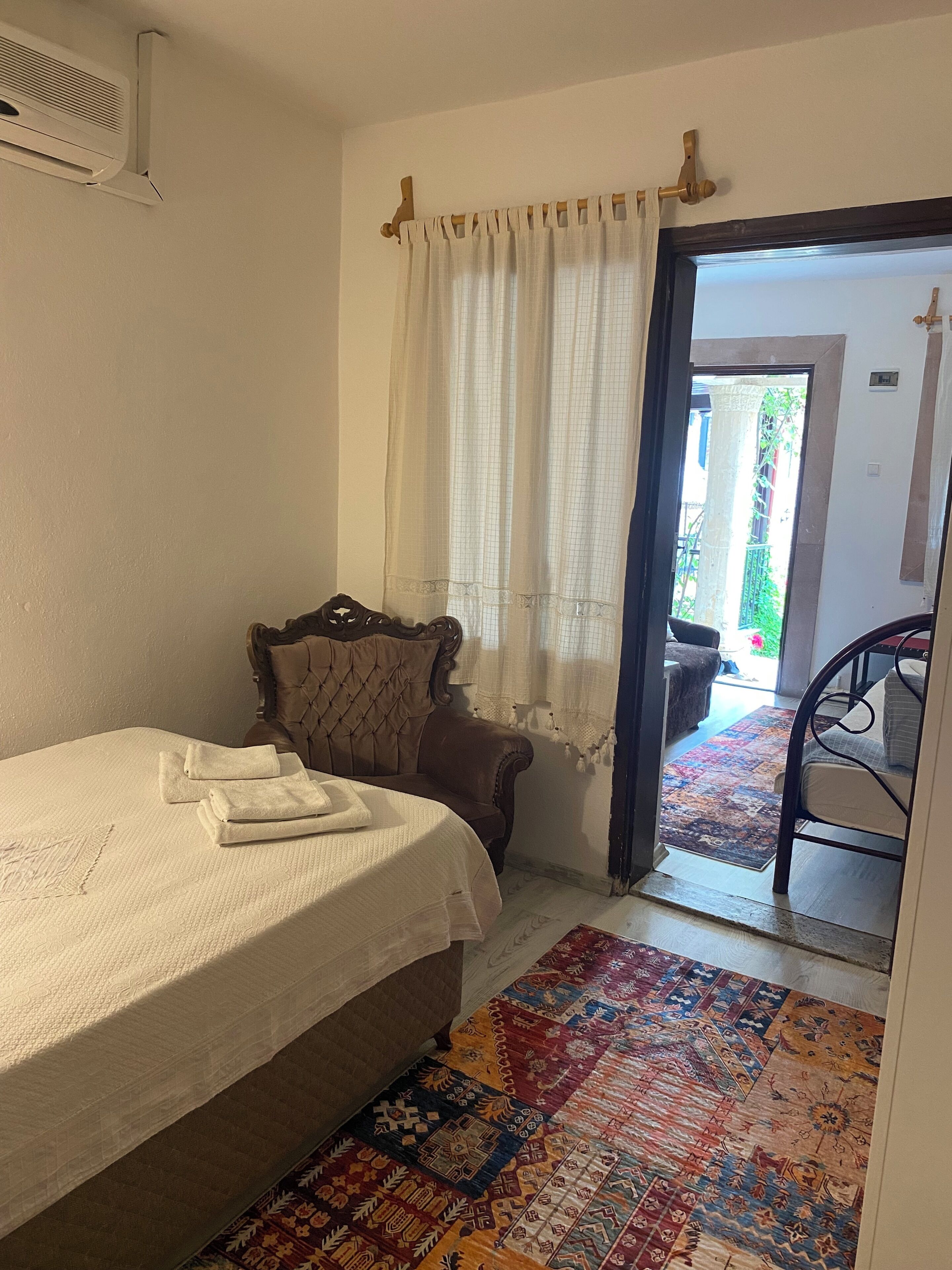 A bedroom with a bed, a brown upholstered chair, a patterned rug, and a view of the outside.