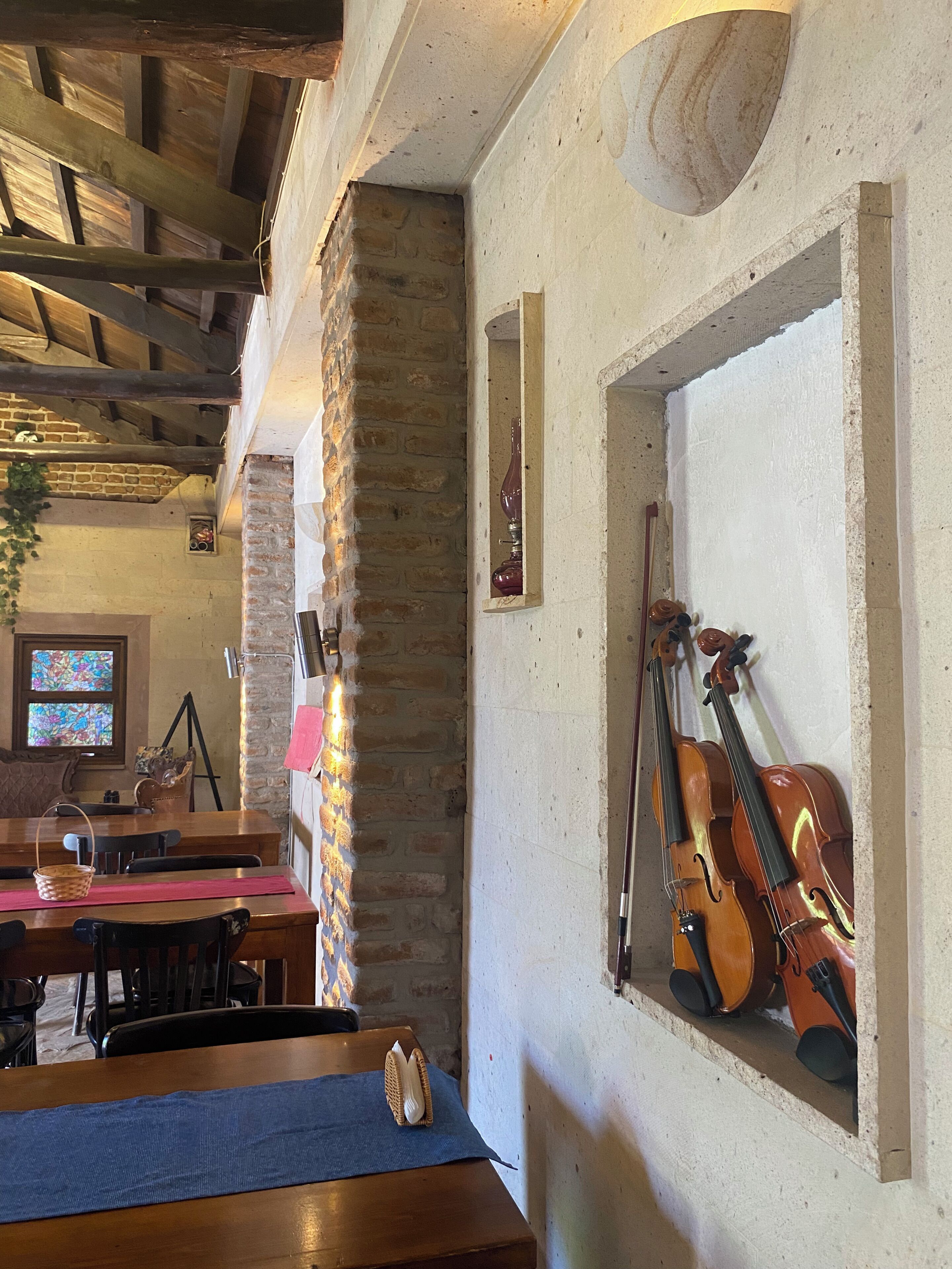 A room with a wooden ceiling, a brick wall, a table with chairs, a violin stand, and a window with stained glass.