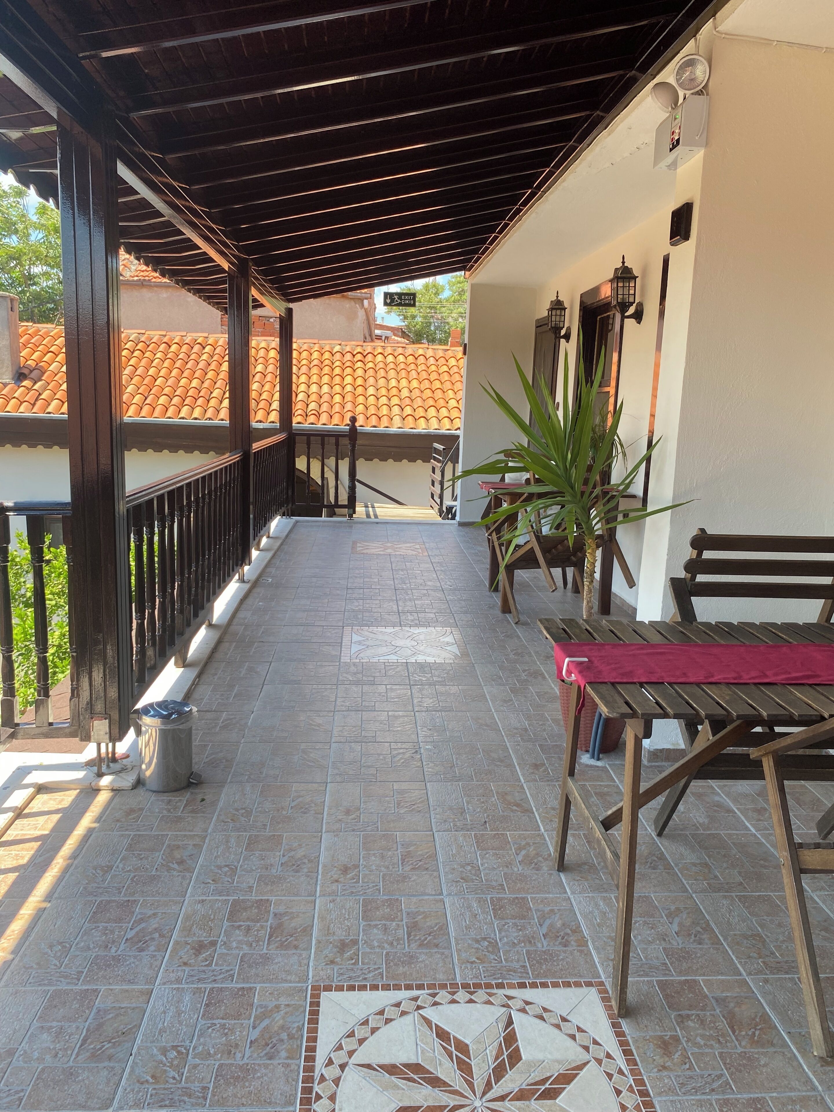 A covered outdoor area with a tiled floor, a bench, a table with a red cloth, and a potted plant.