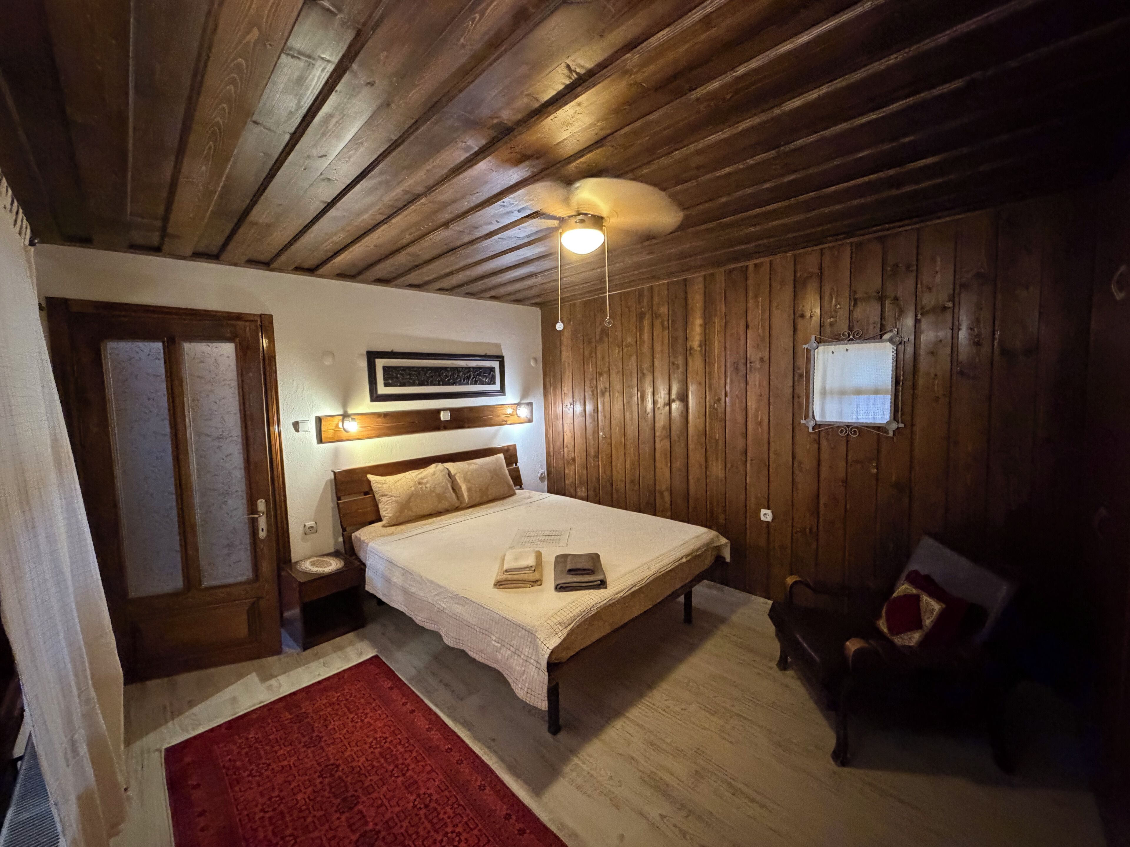 Een kamer met houten lambrisering, een bed, een nachtkastje, een rood tapijt, een stoel en een plafondventilator.