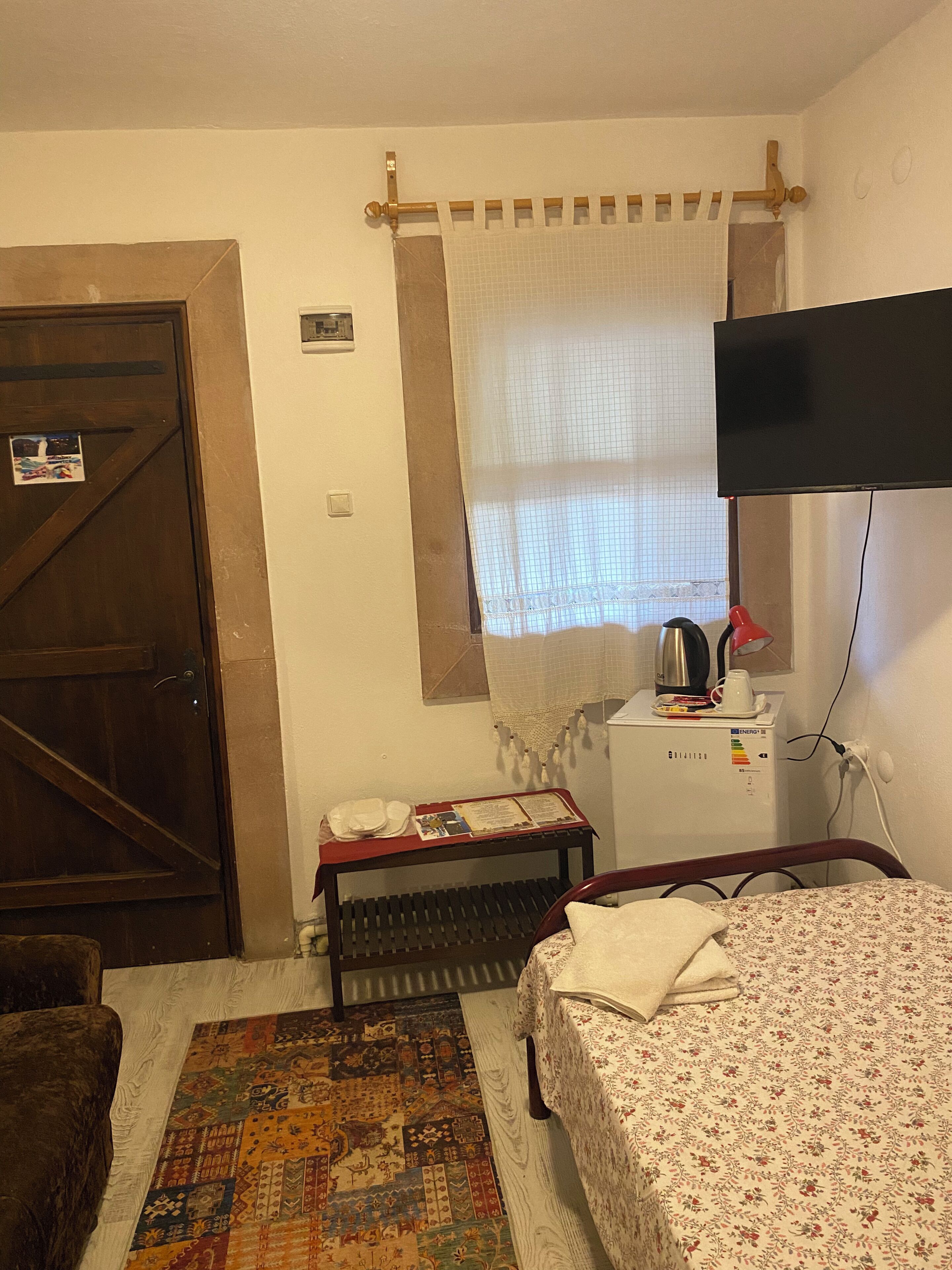 Una habitación con una cama, una puerta de madera, una mesita con una tetera, un televisor y un tapete estampado.