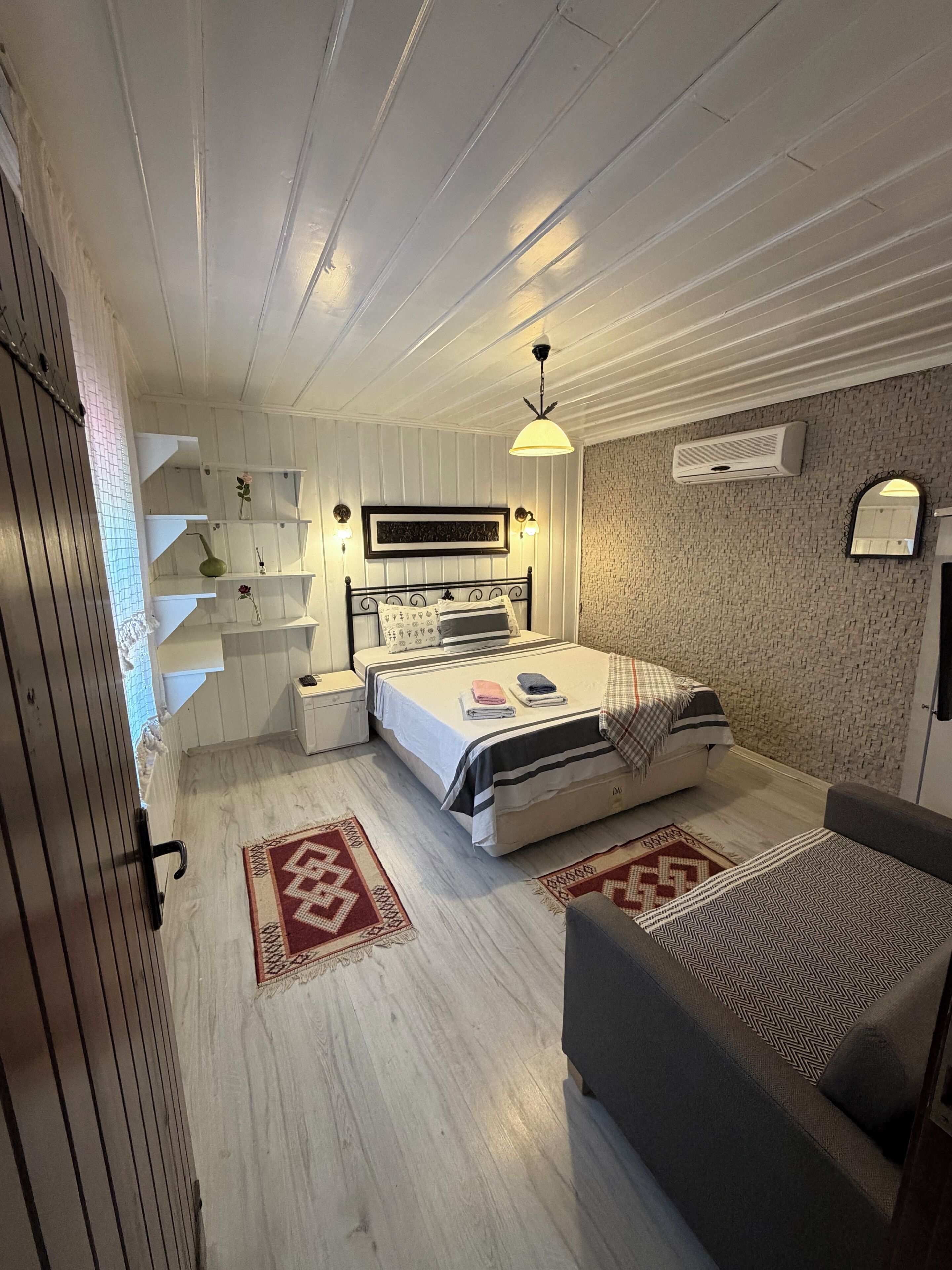 A bedroom with a bed, bedside tables, a hanging light fixture, a wall-mounted air conditioner, and a patterned rug.