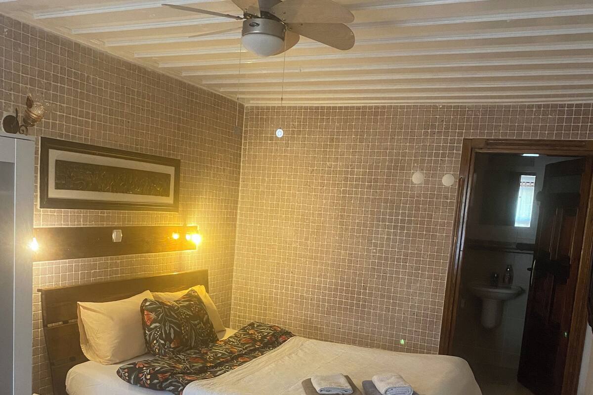 Habitación de hotel con cama, ventilador de techo, mesita de noche, una pequeña mesa y baño visible a través de una puerta abierta.