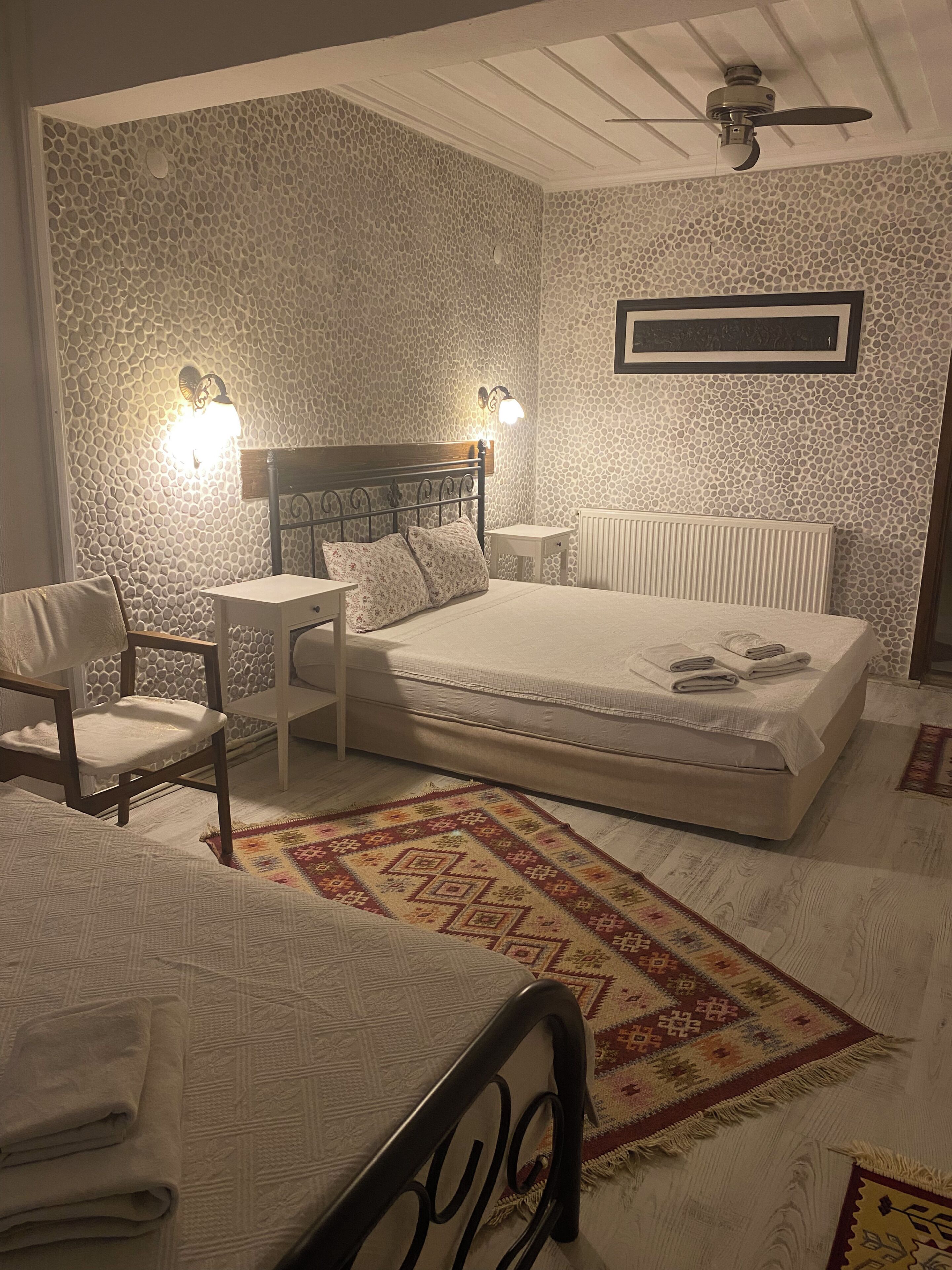 Une chambre à coucher avec un lit, des tables de chevet, une chaise, un tapis à motifs et un ventilateur de plafond.