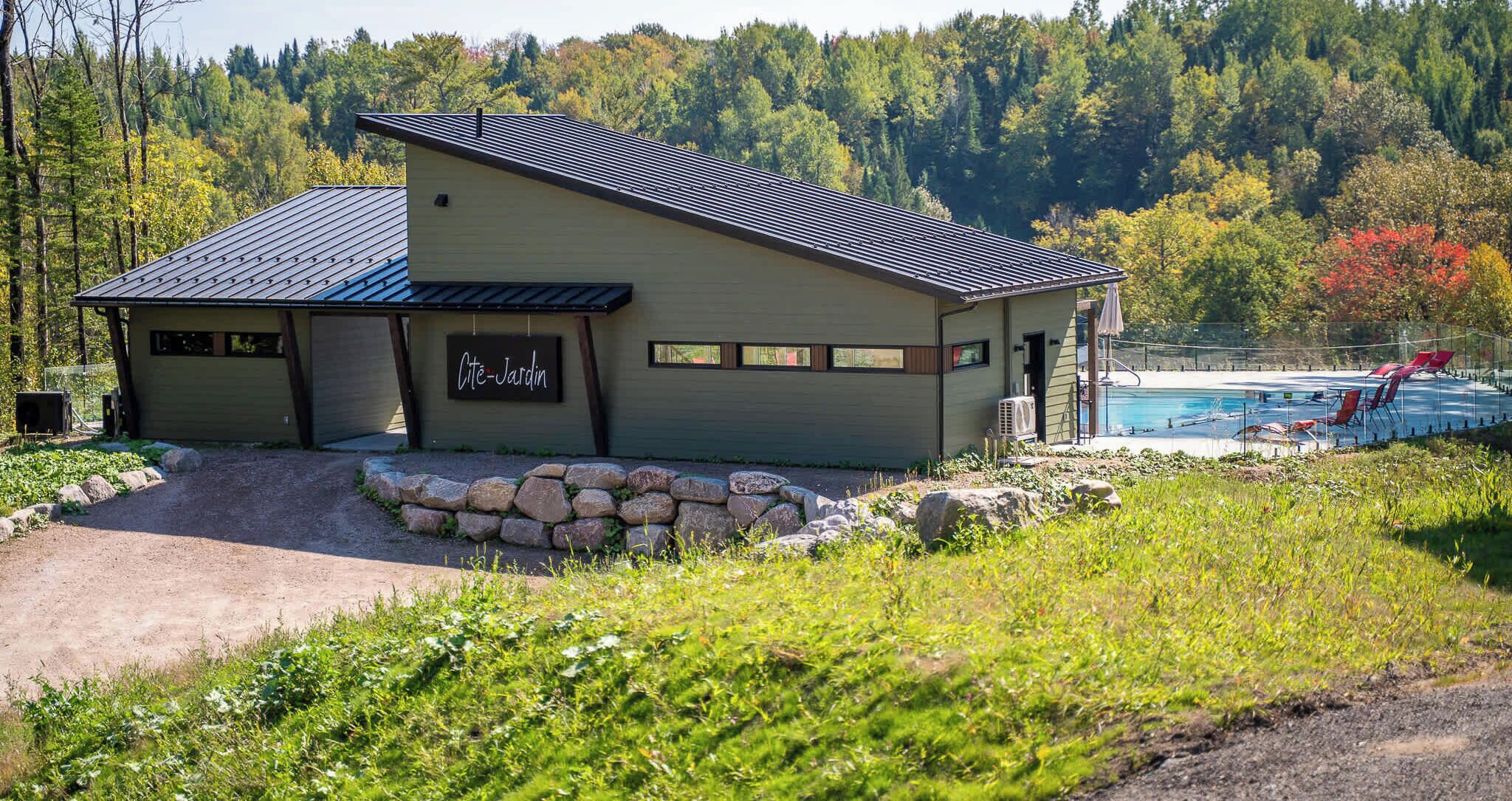 Un chalet avec un chemin de pierre menant à l’entrée, un espace piscine agrémenté de chaises rouges et un décor de forêt en arrière-plan.