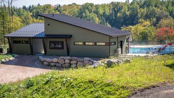 Un chalet avec un chemin de pierre menant à l’entrée, un espace piscine agrémenté de chaises rouges et un décor de forêt en arrière-plan.
