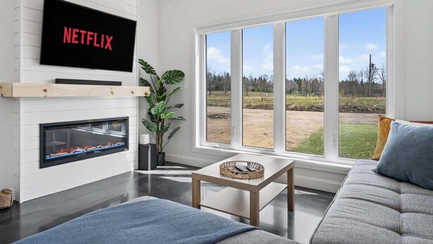 Un salon avec une cheminée, un canapé, une table basse, un téléviseur fixé au mur et une grande fenêtre donnant sur l’extérieur.