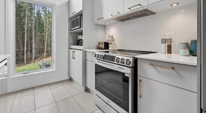 A modern kitchen with white cabinetry, a built-in stove, and a window overlooking a forest.