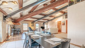 A dining area with a long table set for four, wooden beams on the ceiling, and a television in the background. - Apartment “Maison d’Odette” with Lagoon View, Wi-Fi, and Air Conditioning (Gruissan)