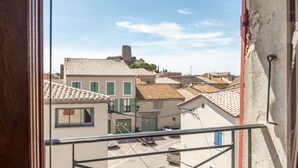 A view from a window overlooking a street with buildings, a tower, and parked cars. - Apartment “Maison d’Odette” with Lagoon View, Wi-Fi, and Air Conditioning (Gruissan)