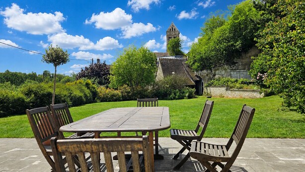 Un patio con una mesa y sillas de madera, un muro de piedra y una torre de iglesia al fondo.